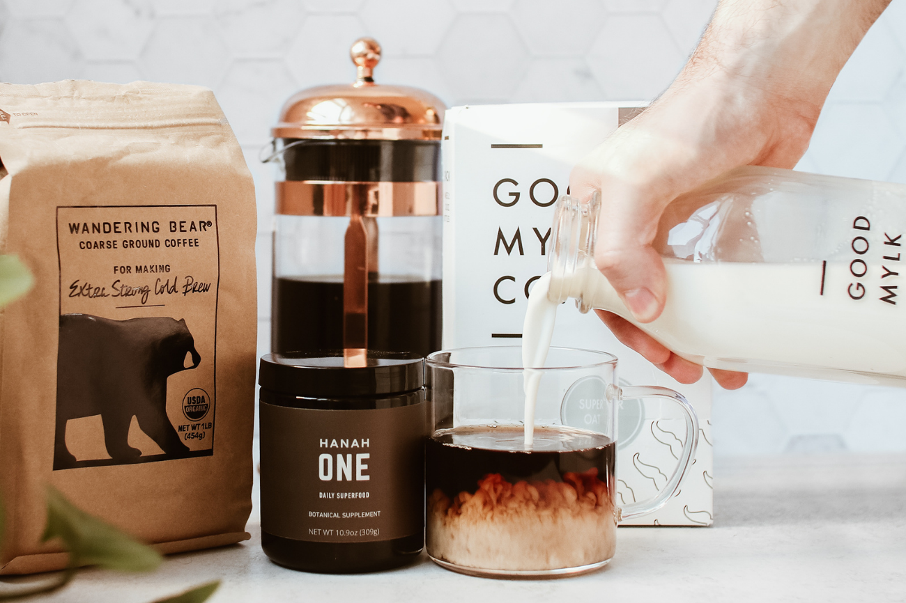 A hand pours Goodmylk Co. oat milk into a glass mug filled with coffee and HANAH ONE.